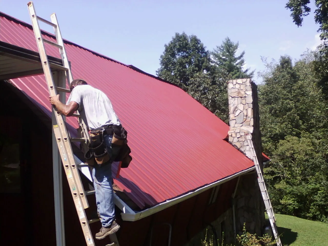 Roofer on a ladder beside a red metal roof and stone chimney — Soffit & Fascia Repair by Roofing Brothers in Falkville
